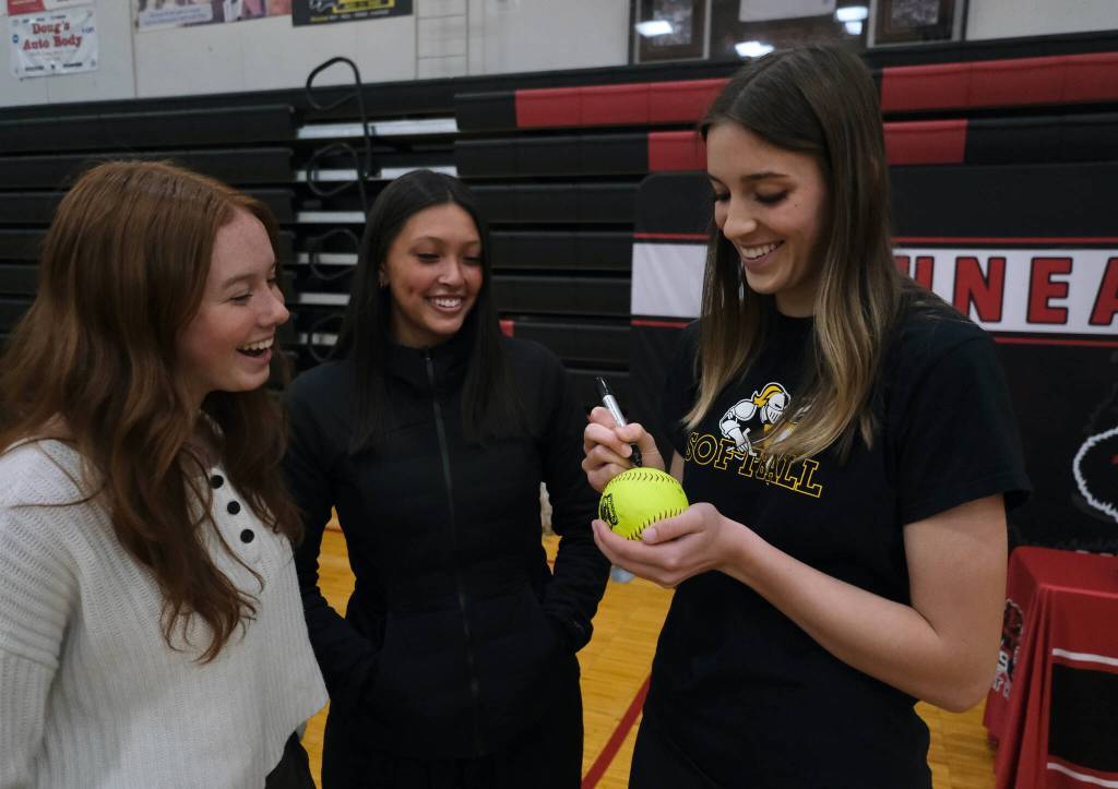 Juneau-Douglas High School: Yadaa.at Kalé senior Mila Hargrave autographs a softball as junior teammate Bristol Casperson and senior teammate Kiah Yadao look on in the George Houston Gymnasium on Monday before Hargrave signed a national letter of intent to attend Pacific Lutheran University and play softball for the Lutes. (Klas Stolpe / For the Juneau Empire)
