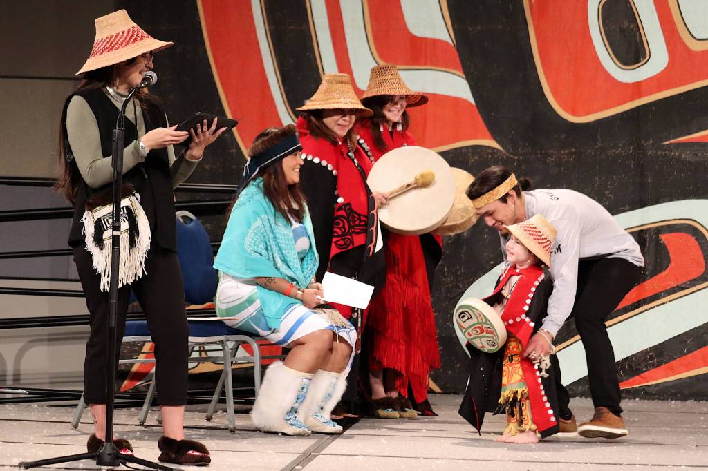 Gyaaaang Peele-Frisby, 2, wears regalia that includes includes a button robe made from the baby blanket of his mother, Sarah Peele, seen at left as she emcees the toddler regalia review as part of Celebration on Thursday at Centennial Hall. (Mark Sabbatini / Juneau Empire)