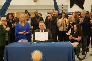 Gov. Mike Dunleavy holds up the omnibus crime bill, House Bill 66, after signing it at a ceremony Thursday at the Department of Public Safetys aircraft hangar at Lake Hood in Anchorage. At his side are Sandy Snodgrass, whose 22-year-old son died in 2021 from a fentanyl overdose, and Angela Harris, who was stabbed in 2022 by a mentally disturbed man at the public library in Anchorage and injured so badly that she now uses a wheelchair. Snodgrass and Harris advocated for provisions in the bill.Behind them are legislators, law enforcement officers and others. (Yereth Rosen/Alaska Beacon)