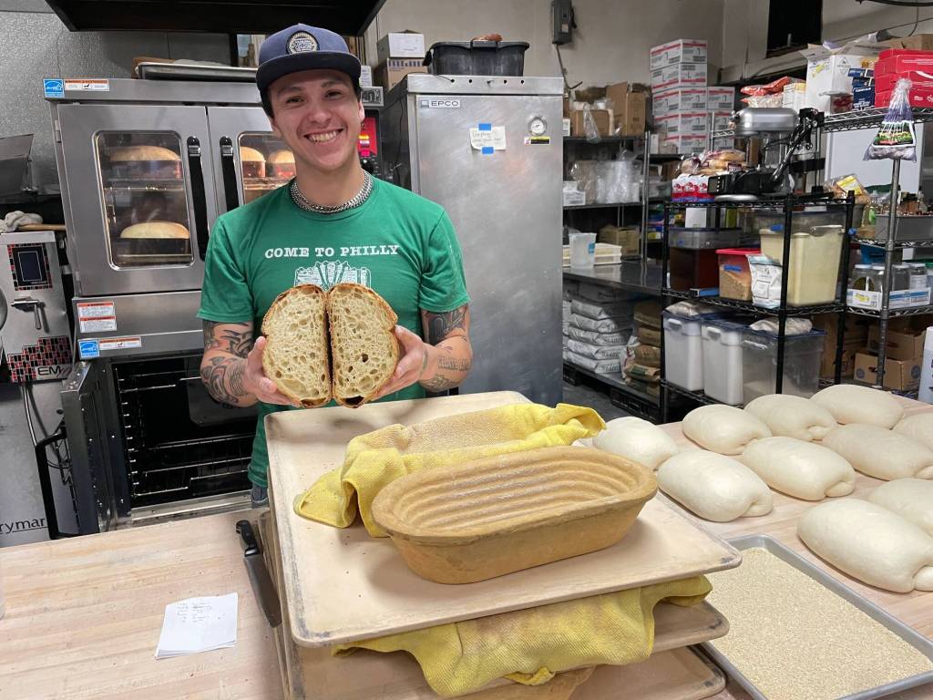 Josh Druley, known as the DoughFather, with a freshly sliced loaf of his sourdough bread baked in the new stacked Vulcan ovens seen behind him in the kitchen at In Bocca Al Lupo. Rising bread dough mounds rest on the table beside him. (Laurie Craig / Juneau Empire)