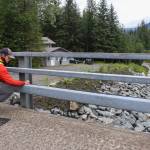 Jonathan Tuttle, a hydrologist with the U.S. Geological Survey, installs monitoring equipment at the Mendenhall River bridge on Back Loop Road Thursday morning. Beyond the bridge, a Juneau resident living along the Mendenhall River looks out at the water. (Jasz Garrett / Juneau Empire)