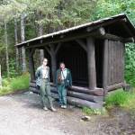 U.S. Forest Service park rangers Rachael Rosenbaum and Isabel Dziak stand beside the Civilian Conservation Corps log shelter on the Trail of Time in June of 2024. The shelter was initially built when the glacier was visible from this site. (Laurie Craig / Juneau Empire)