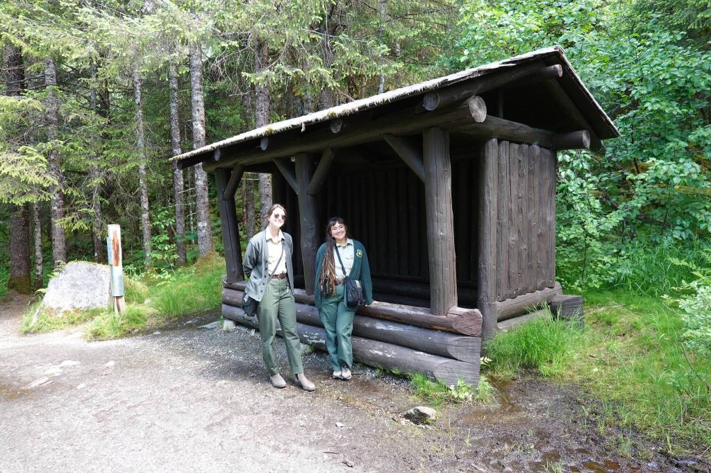 U.S. Forest Service park rangers Rachael Rosenbaum and Isabel Dziak stand beside the Civilian Conservation Corps log shelter on the Trail of Time in June of 2024. The shelter was initially built when the glacier was visible from this site. (Laurie Craig / Juneau Empire)