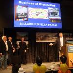 Mitch Falk (right), owner of Bullwinkles Pizza Parlor, offers a tribute to Bill Adair (far left on stage), the original owner who founded the company in 1973, as the pizza establishment was named the Greater Juneau Chamber of Commerces Business of Distinction for 2024 on Saturday at Centennial Hall. (Mark Sabbatini / Juneau Empire)