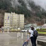 Aurelie Alexander, with her young son Robin in his stroller, photographs a helicopter hoisting cellular equipment onto the roof of the Marine View building at midday Wednesday. As a resident of the apartment/office building, she and others were notified to leave the building during the helicopter operation.(Laurie Craig / Juneau Empire)