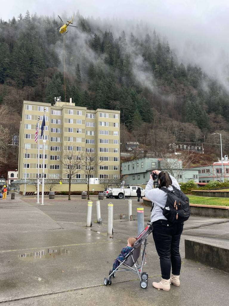 Aurelie Alexander, with her young son Robin in his stroller, photographs a helicopter hoisting cellular equipment onto the roof of the Marine View building at midday Wednesday. As a resident of the apartment/office building, she and others were notified to leave the building during the helicopter operation.(Laurie Craig / Juneau Empire)