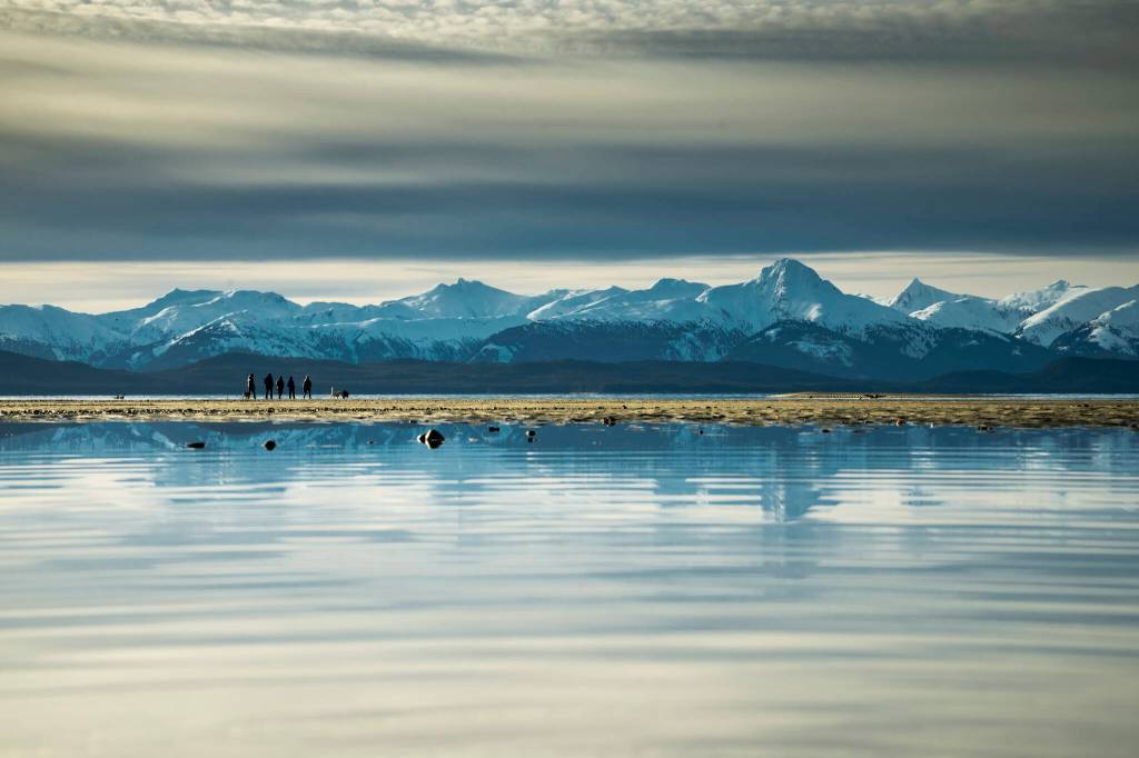 Dogs and their owners walk on Eagle Beach, with the snowcapped Chilkat mountain range rambling jaggedly across the horizon, in Juneau, Alaska, Jan. 29, 2023. The states capital, a popular cruise port in summer, becomes a bargain-seekers base for skiing, skating, hiking and glacier-gazing in the winter off-season. (Christopher S. Miller/The New York Times)