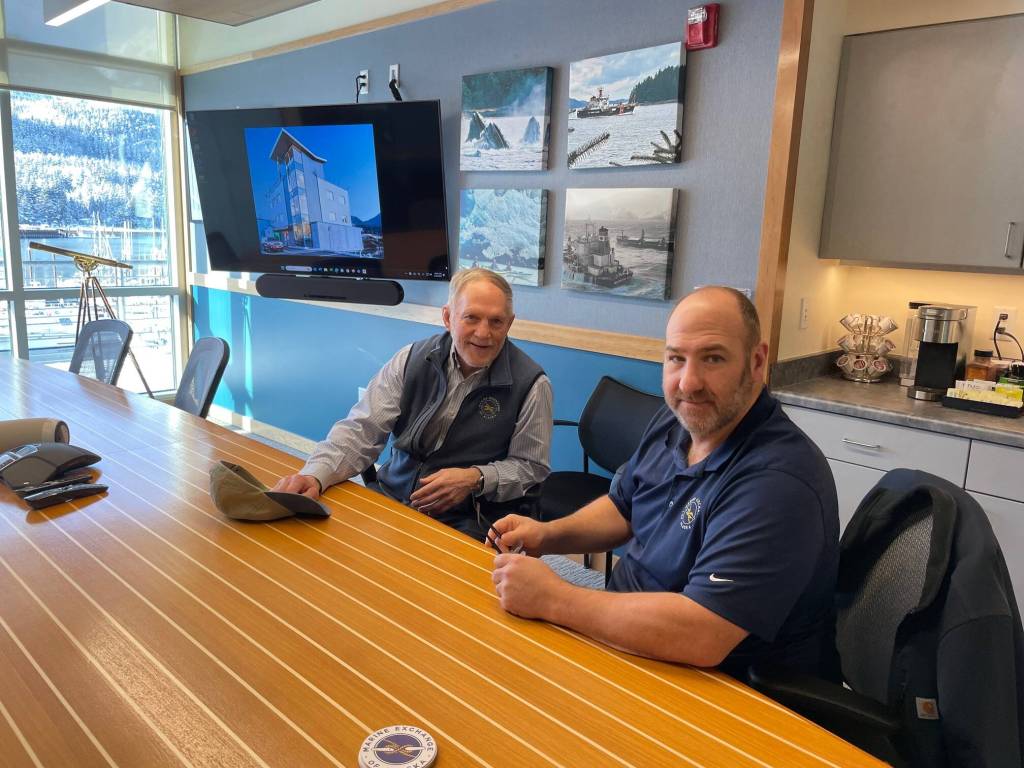 Ed Page and Steve White talk at MXAKs ship-deck designed conference table with a classic long glass on a brass tripod by the window. On the wall between the men is a photo of the wave-splashed ship Selendang Ayu, a memorable disaster. (Laurie Craig / Juneau Empire)