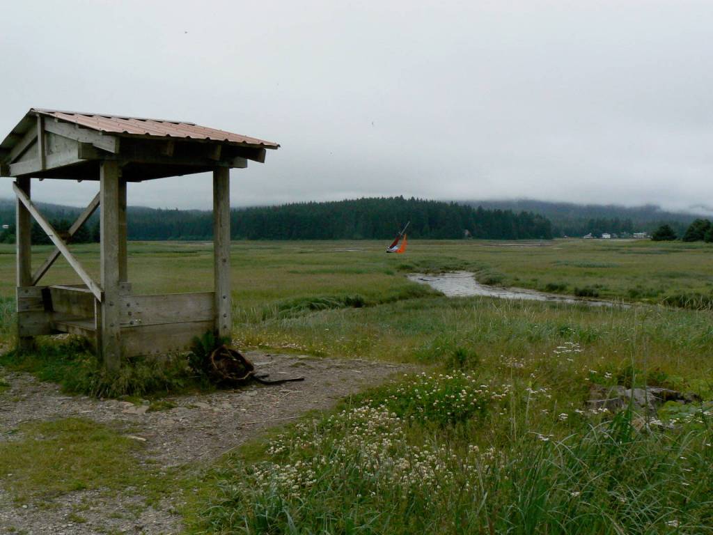 A rusty wheel, remnant of old automobiles used decades ago to reinforce the dike, leans against Corey McKrills Eagle Scout covered bench in this 2007 photo. River otters lived in an old DeSoto in a dredge pond where they ran across the trail or swam nearby to catch flounder. Red sails on a boat in the distance show where a sailor misjudged the Gastineau Channel tide and ran aground. (Photo by Laurie Craig)