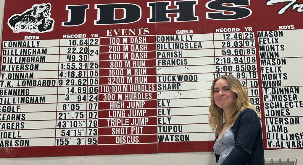 Bella Connally, who just finished her Juneau-Douglas High School: Yadaa.at Kalé freshman track and field season is shown next to her school record for the 100 meters on Wednesday at the JDHS auxiliary gym. (Photo courtesy Chris Connally)