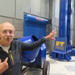 Billy Connor gestures toward load frames  the large blue beams behind him  in the Usibelli Buildings high bay at the University of Alaska Fairbanks in July 2026. (UAF photo by Sam Bishop)